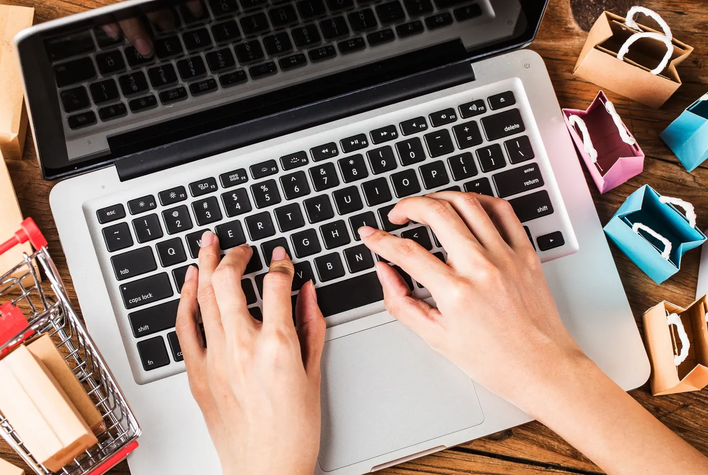 An overhead view of a laptop, miniature shopping trolley and paper bags representing online shopping cues