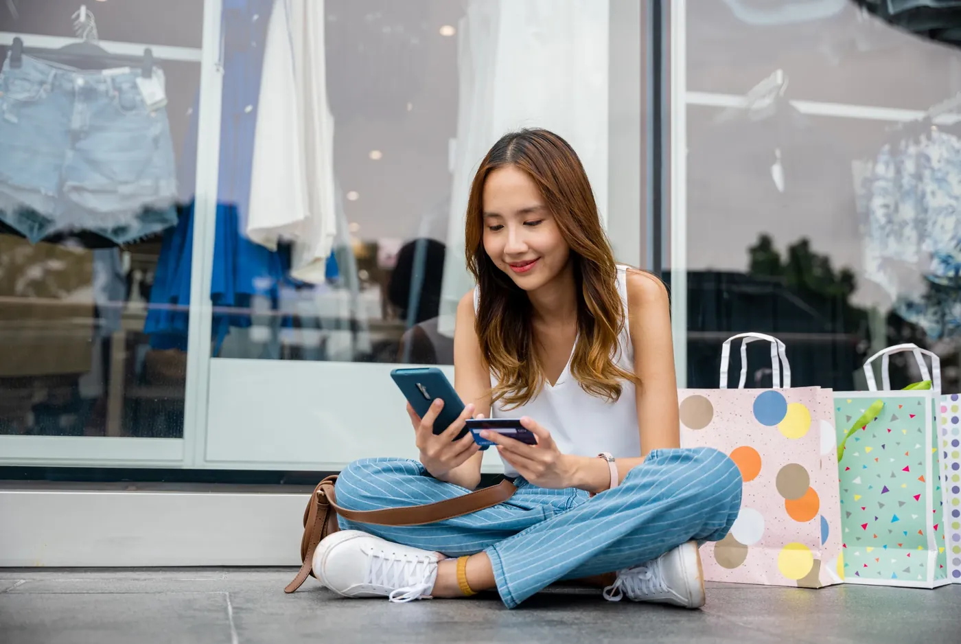 A person seated outside a shop checking a phone and holding a payment card